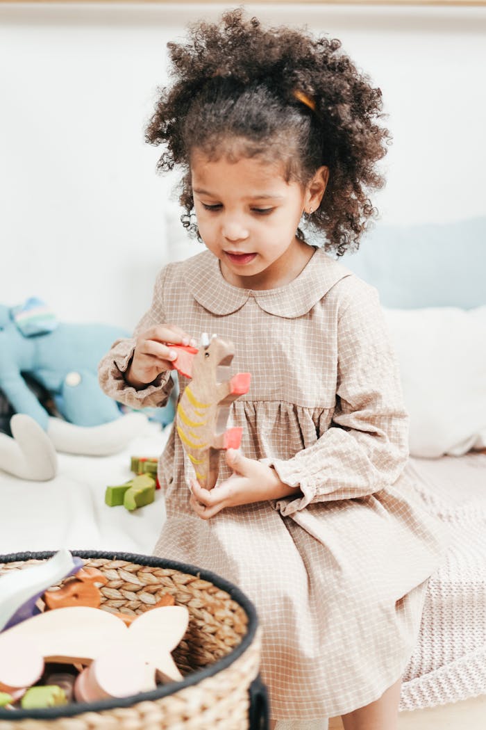Menu Young girl with afro-textured hair enjoying playtime with wooden toys in a cozy bedroom setting.